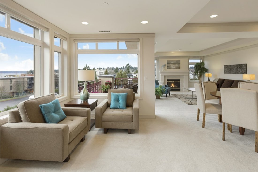 Real estate photography of a bright, coastal-modern living room with large windows and teal accent chairs.